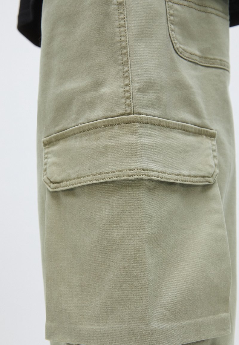 Khakifarbene Cargo-Shorts aus einem strukturierten Baumwollgemisch. Sie verfügen über eine Seitentasche mit Klappe und sichtbare Nähte.