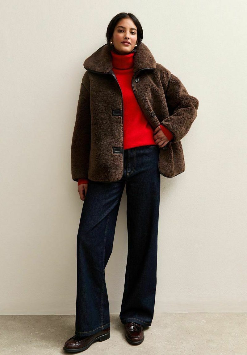 Brązowa kurtka z futra sztucznego z szerokim kołnierzem i czarnymi guzikami; czerwony golf, ciemne dżinsowe spodnie z szerokimi nogawkami oraz ciemne buty dopełniają całość.