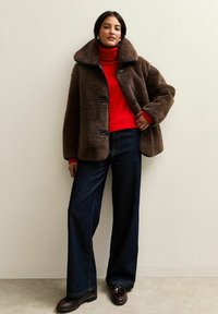 Giacca in pelliccia sintetica marrone con ampio colletto, bottoni neri; dolcevita rosso, pantaloni a gamba larga in denim scuro e scarpe scure completano l'outfit.