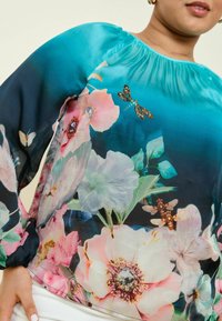 Blusa con stampa floreale in turchese con maniche lunghe a sbuffo. Presenta vari fiori, farfalle e libellule con una texture setosa e scollo arricciato.