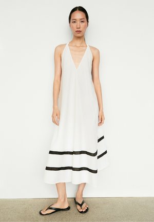 Femme portant une robe midi blanche sans manches avec des rayures horizontales noires près de l'ourlet et des sandales plates noires, debout contre un mur blanc.