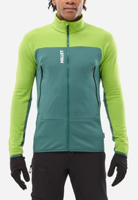 Man wearing a green and teal full-zip jacket with "MILLET" logo, black gloves, and black pants, standing against a white background.