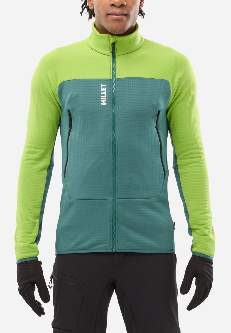 Man wearing a green and teal full-zip jacket with "MILLET" logo, black gloves, and black pants, standing against a white background.