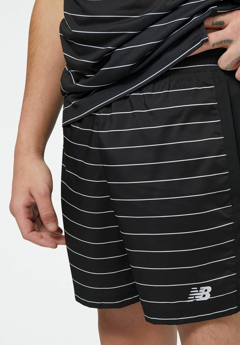 Mann in schwarzen Sportshorts und passendem Shirt mit dünnen weißen Horizontalstreifen, auf den Shorts ist das New Balance-Logo zu sehen.