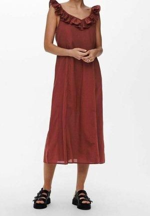 Bezrekawowa sukienka midi w kolorze rdzy z falbankowym dekoltem, noszona z czarnymi sandałami z klamrą, prezentowana na modelce na białym tle.