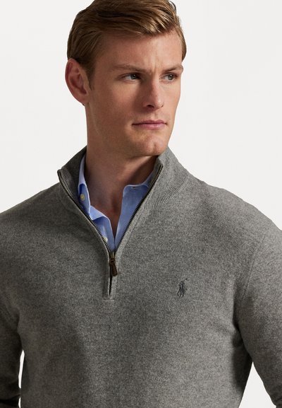 Grå strikket sweater med lynlås krave, der har en kontrasterende indvendig blå skjorte. Bemærkelsesværdig logo detalje på brystet, glat tekstur.