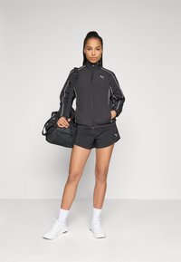 Mulher com uma jaqueta desportiva preta da Puma e calções, sapatilhas brancas, segurando uma bolsa desportiva preta sobre o ombro, em frente a um fundo liso.