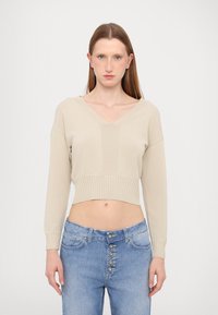 Maglione beige corto con maniche lunghe e scollo a V, caratterizzato da orlo a coste e texture uniforme. Abbinato a jeans blu a vita alta con chiusura a bottoni.
