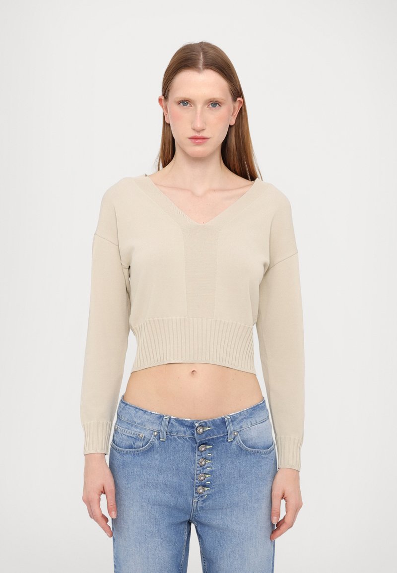 Maglione beige corto con maniche lunghe e scollo a V, caratterizzato da orlo a coste e texture uniforme. Abbinato a jeans blu a vita alta con chiusura a bottoni.