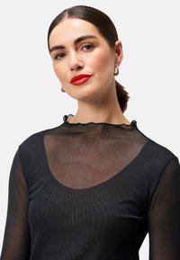 Haut noir transparent à manches longues avec un col rond. Présente une finition texturée subtile et un bord ruffled au niveau du col.