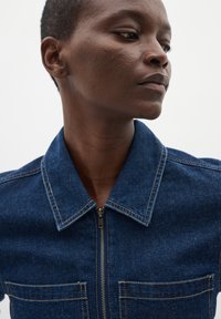 Donkerblauwe denimjack met een rits aan de voorkant, puntige kraag en twee borstzakken. Heeft contrasterende stiksels. Soepele textuur.