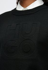 Maglione nero lavorato a maglia con logo "HUGO" tonale e testurizzato sul davanti. Design con scollo rotondo, polsini e orlo a coste.