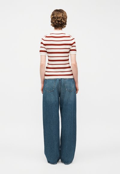 Personne aux cheveux courts et bouclés portant une chemise à manches courtes rayée blanche et rouge et un jean bleu à jambes larges, debout dos au spectateur.