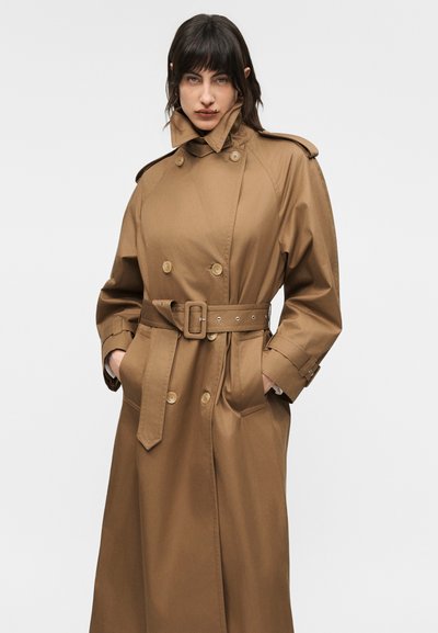 Γυναίκα που φοράει μακρύ διπλό παλτό καφέ ανοιχτής απόχρωσης με ζώνη και σηκωμένο γιακά, έχει τα χέρια στις τσέπες και στέκεται μπροστά σε ένα απλό ανοιχτόχρωμο φόντο.