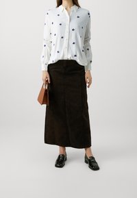 Blusa bianca con ricami floreali blu, chiusura a bottoni sul davanti, maniche lunghe, abbinata a una gonna maxi in denim marrone scuro. Mocassini neri e una borsa marrone.
