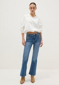 Sudadera blanca recortada con mangas extragrandes, combinada con jeans azules acampanados con bolsillos tachonados. Botines de tacón color canela completan el conjunto.