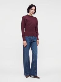 Femme portant un haut bordeaux à manches longues, un jean bleu à jambes larges et des chaussures brunes pointues, debout devant un fond blanc uni.