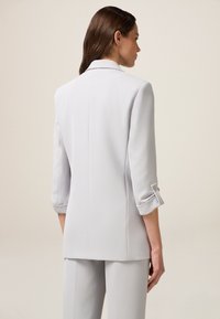 Light gray tailored blazer with a structured design, three-quarter sleeves, and cuffs featuring buttons. Smooth fabric texture, minimalistic style.