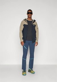 Vyras, dėvintis juodą ir smėlio spalvos užtrauktuku užsegamą striukę, juodą marškinėlį, mėlynas kelnes su turkio spalvos akcentais, žalias ir mėlynas batus, bei juodus akinius nuo saulės, stovi.