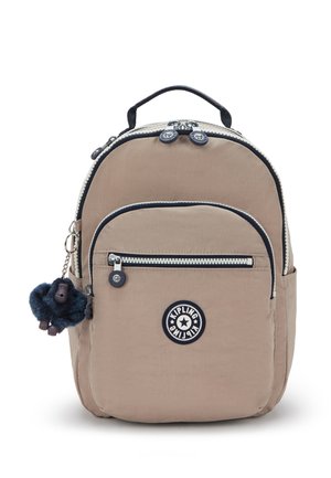 Beige Kipling-rugzak met voorvak met rits, zwarte bies, bovenhandvat en een kleine pluchen apensleutelhanger aan de zijkant.