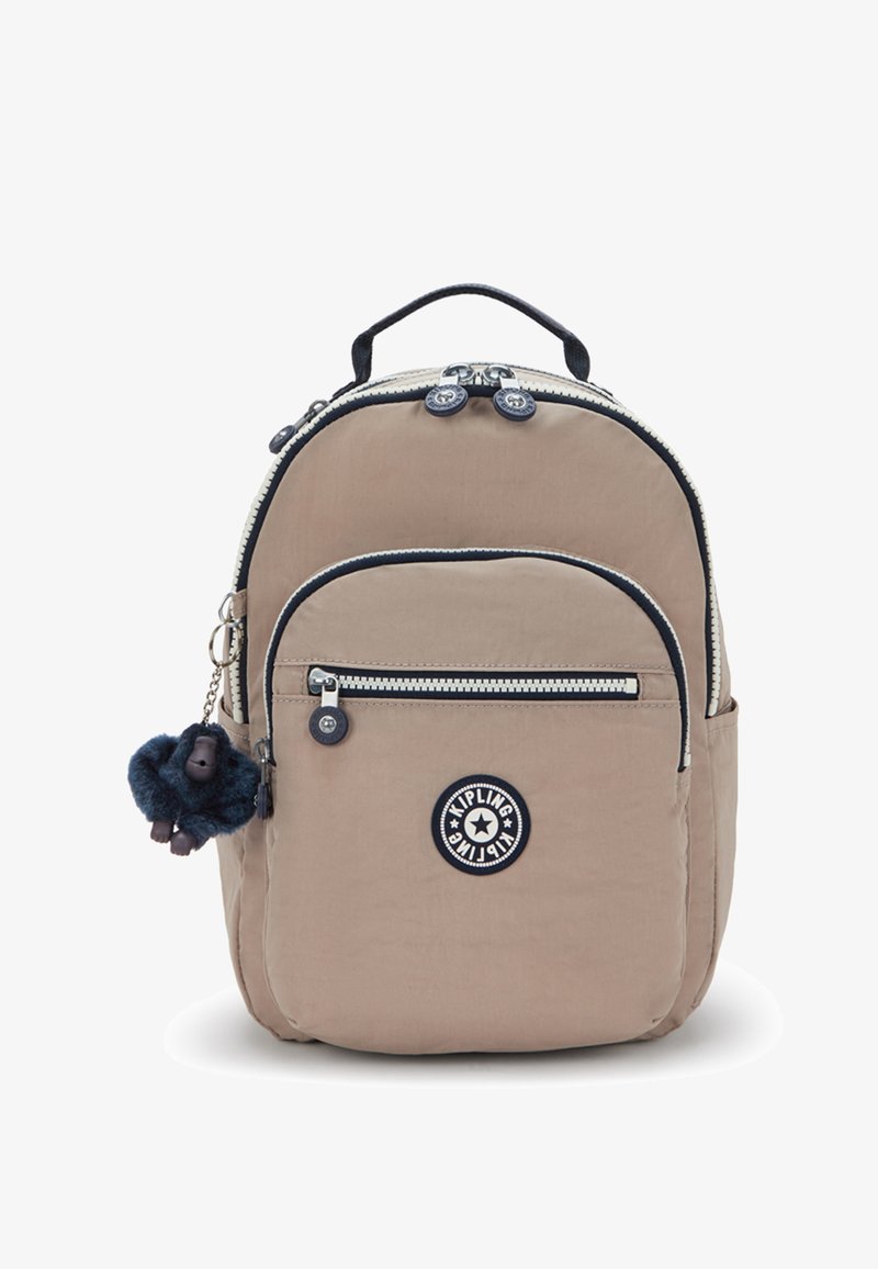 Beige Kipling-reppu, jossa edessä vetoketjutasku, mustat reunukset, yläkahva ja pieni pehmeä apina-avainperä sivussa kiinnitettynä.
