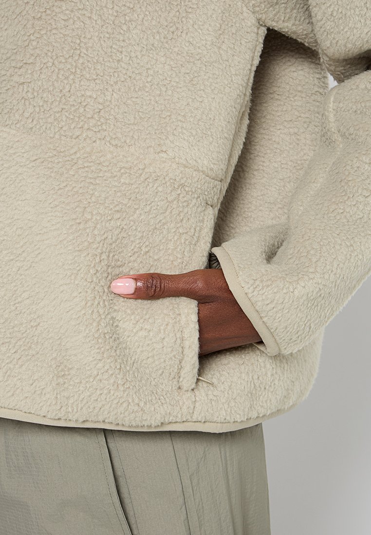 Veste en polaire beige avec une surface texturée et une poche kangourou, dotée d'un ourlet et de poignets en tissu lisse contrastants.