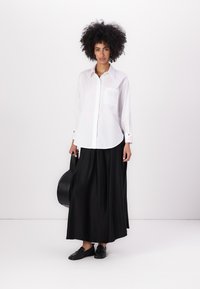 White long-sleeve shirt with a pocket, paired with a flowing black maxi skirt. Black shoes and a black bag complete the outfit.