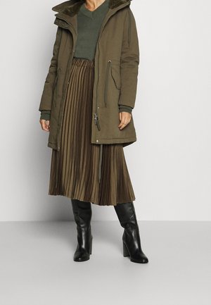Femme portant une parka vert olive, une jupe midi plissée, un pull assorti et des bottes noires à talons montant jusqu'aux genoux, debout sur un fond uni.