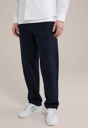Jūriniai chinos, pagaminti iš minkštos medžiagos, su tiesiomis kojomis ir priekinėmis kišenėmis. Dėvimi su baltais sportiniais batais, ant kurių matomas logotipas.