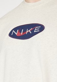 Närbild på en vit skjorta med en blå oval Nike-logotyp i form av en tygmärke på bröstet, med en röd swoosh och vita broderade bokstäver.