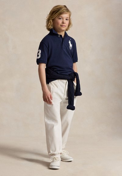 Vaaleahiuksinen lapsi pukeutuneena tummansiniseen poolopaitaan, valkoisiin housuihin ja valkoisiin tennareihin, tummansininen neule sidottuna vyötärölle, seisoo neutraalia taustaa vasten.