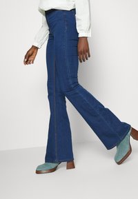 Uppflutna jeans i mörkblått med en slät textur. Har synliga sid sömmar och fickkonstruktion. Matchas med gröna klackskor.