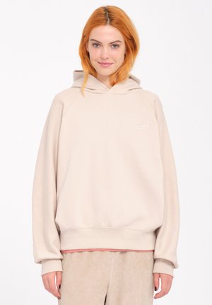 Sweat-shirt à capuche beige avec une coupe décontractée, présentant des poignets et un ourlet côtelés. Petit logo sur la poitrine, porté par-dessus un haut rose et un pantalon en velours côtelé.
