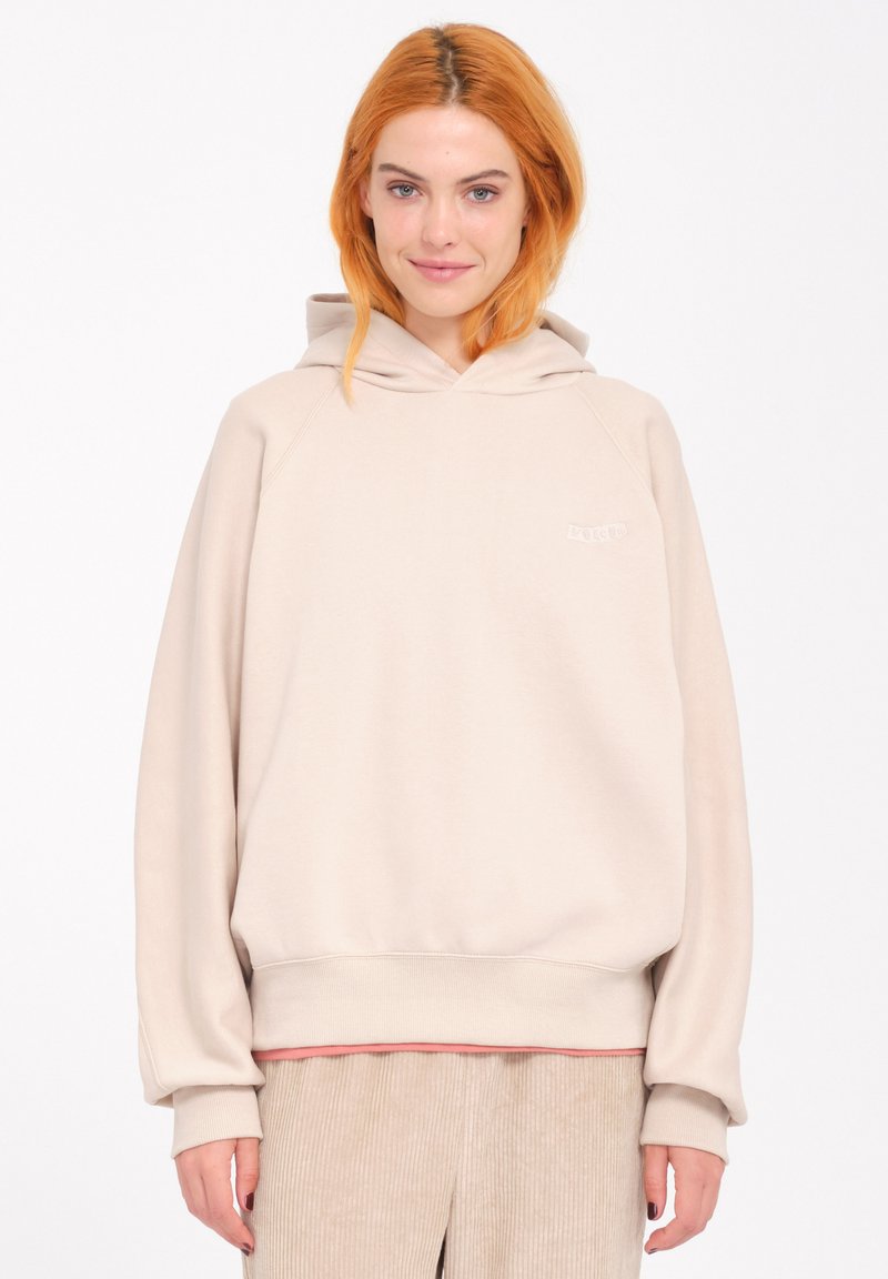 Beige hooded sweatshirt with a relaxed fit, featuring ribbed cuffs and hem. Small logo on the chest, worn over a pink top and corduroy pants.