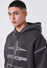 Sudadera con cremallera en color gris, con capucha, que presenta costuras blancas y detalles del logo. Textura suave con un corte relajado, diseñada para mayor comodidad.