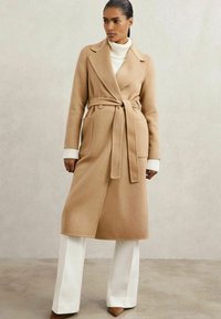 Abrigo trench de mezcla de lana en color marrón claro con cinturón, solapas anchas, bolsillos frontales y un ajuste entallado, combinado con un suéter de cuello alto en color crema y pantalones blancos.