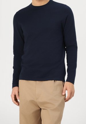 Mand iført en tætsiddende marineblå langærmet skjorte og beige bukser, stående mod en ensfarvet lys baggrund.