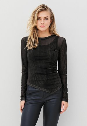 Schwarzes langärmliges Oberteil aus transparentem Stoff mit Rundhalsausschnitt und asymmetrischem Saum, kombiniert mit enganliegenden dunklen Hosen.