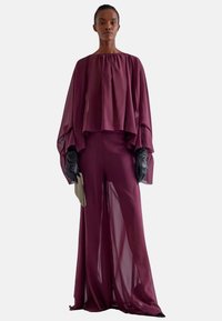 Completo bordeaux a due pezzi con un top fluttuante a strati e pantaloni ampi e trasparenti. Guanti neri e una pochette slim completano il look.