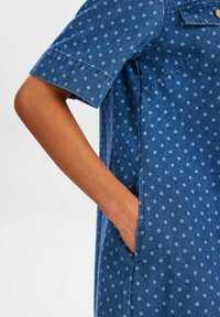 Robe en denim bleu foncé avec des pois bleu clair, dotée de manches courtes et d'une poche sur le côté. Texture de tissu structurée.