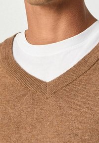 Gros plan d'une personne portant un t-shirt blanc à col rond sous un pull en maille marron à col en V.