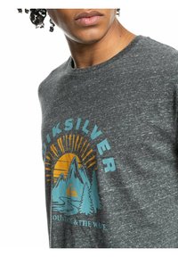 Primer plano de una persona que lleva una camiseta gris oscuro de Quiksilver con un gráfico de montaña, sol y árbol, y el texto "Montaña y La Ola".