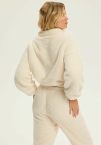 Kurtka z futra sztucznego w kolorze kremowym, z wysokim kołnierzem, oversize'owymi rękawami i elastycznym pasem, prezentowana z tyłu.