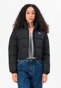 Black cropped puffer jacket with a high collar, quilted design, zip closure, and logo detail on the chest. Paired with blue high-waisted jeans.