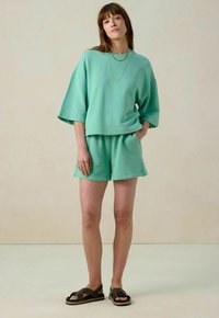 Sweatshirt ample en mélange de coton vert clair associé à un short assorti. Comprend une coupe décontractée et des manches 3/4. Des sandales plates complètent le look.