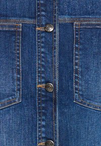 Denim odjevni predmet tamnoplave boje, sa srebrnim metalnim dugmadima, vidljivim šavovima i dva prednja džepa.
