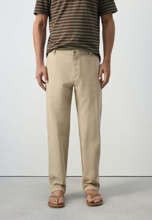 Mann trägt beige gerade Hose, kombiniert mit braunen Flip-Flop-Sandalen und einem braun gestreiften kurzärmligen Hemd.