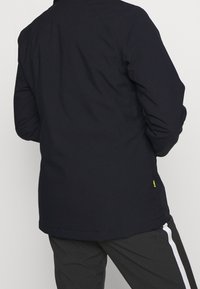 Giacca dark navy con tessuto liscio, design aderente e sottile accento del logo giallo. Maniche lunghe e orlo posteriore allungato.