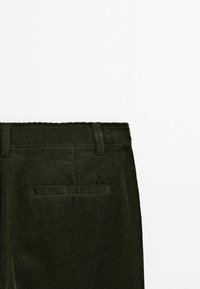 Pantalon en velours côtelé vert foncé avec poche passepoilée à l'arrière et passants de ceinture sur fond blanc.