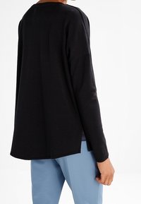 Osoba oblečená v volném černém dlouhém rukávu a světle modrých kalhotách, zobrazená zezadu na bílém pozadí.